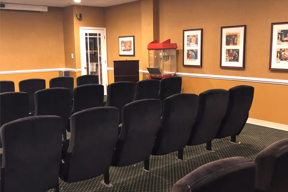 A small theater or presentation room with rows of black cushioned chairs facing a podium and a popcorn machine against a mustard yellow wall. Several framed pictures hang on the wall, and there is a white door with glass panels in the background.