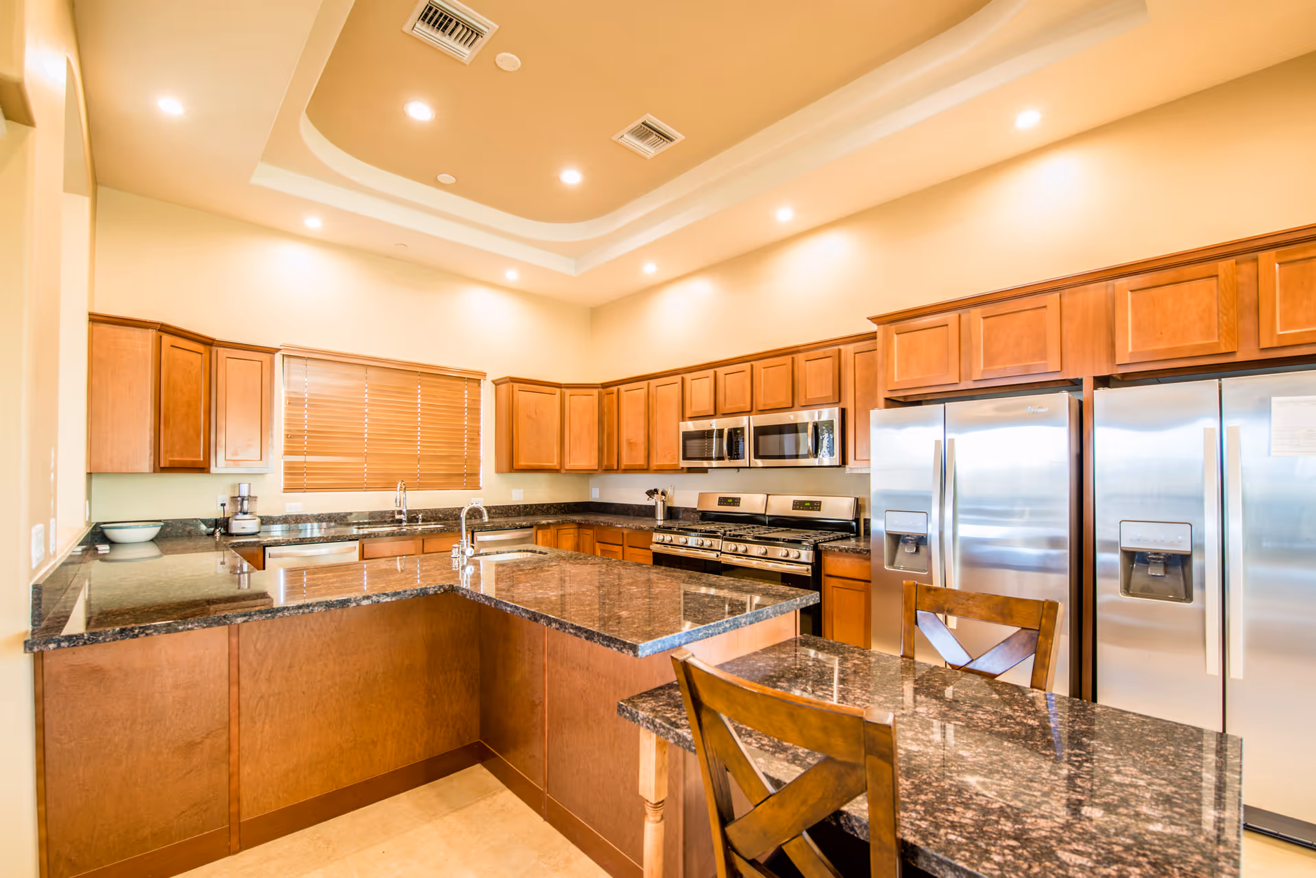 Spacious kitchen with a large granite island, wooden cabinets, stainless steel refrigerators and range, and recessed ceiling lights.