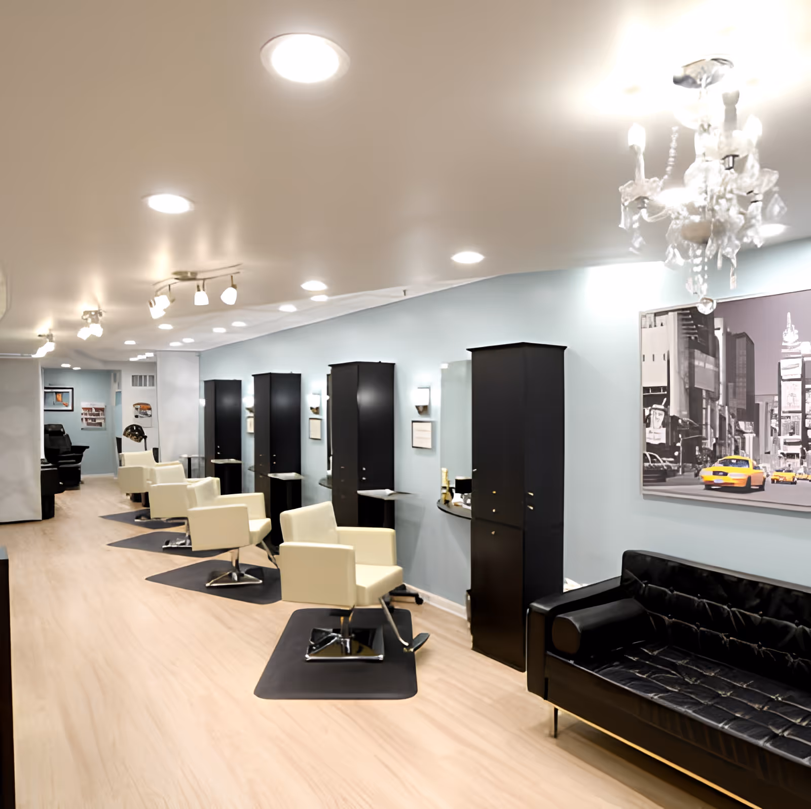 Interior view of a modern salon area with multiple white styling chairs in front of mirrors and black storage cabinets. The floor is light wood, and the walls are painted light blue. There is a black leather couch on the right side under a large black and white cityscape photo with yellow taxis. The ceiling has recessed lighting and a decorative chandelier.