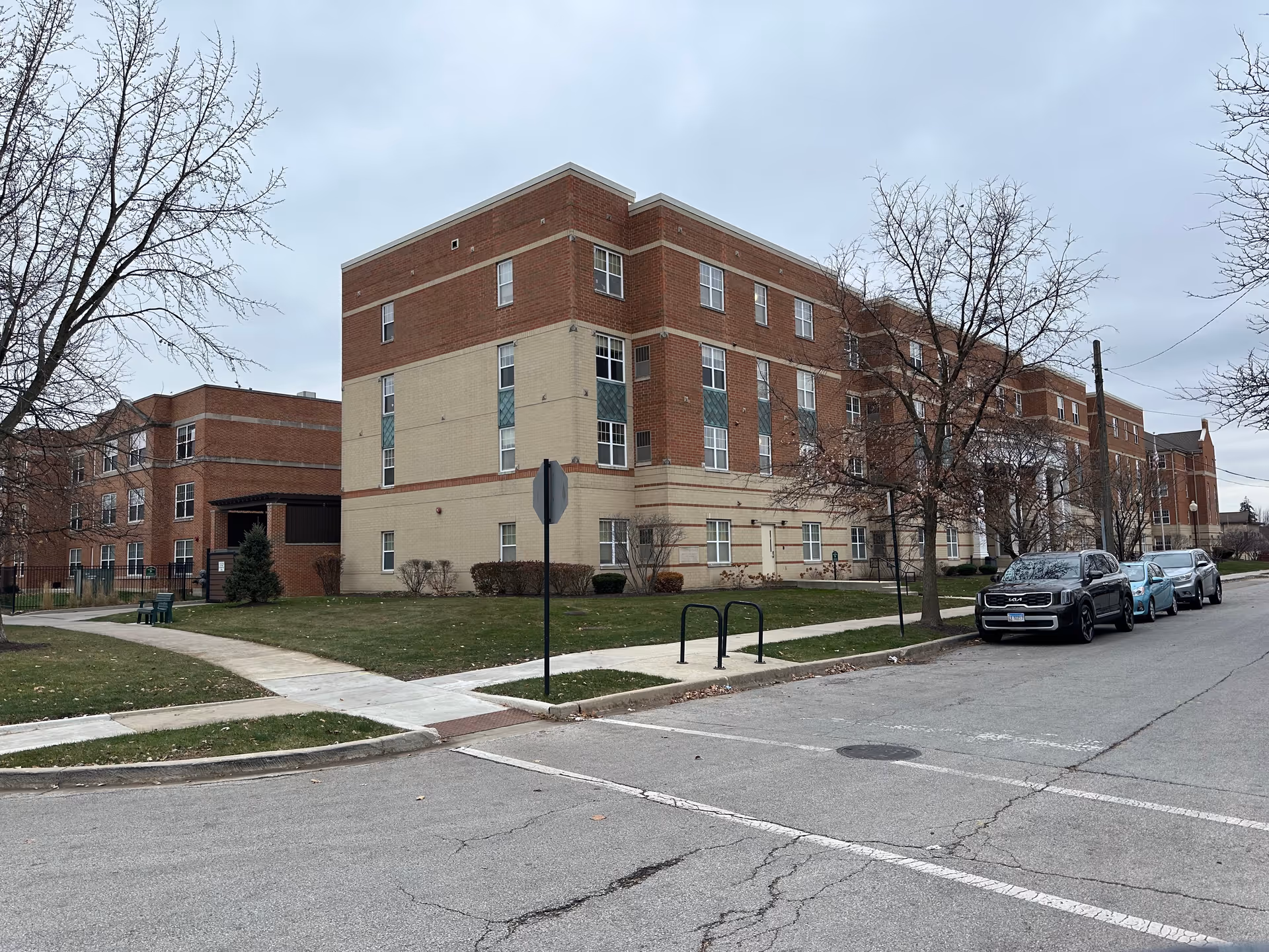 Brick multi-story senior living building on a street corner with parked cars and leafless trees.