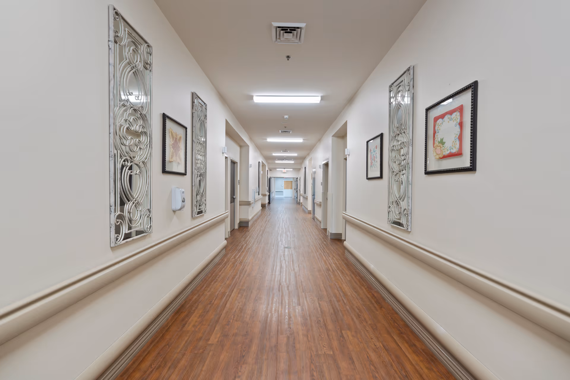 A long, well-lit hallway in a senior living facility with wooden flooring, beige walls, decorative wall art, and handrails on both sides. Several doorways line the corridor, and ceiling lights illuminate the space.