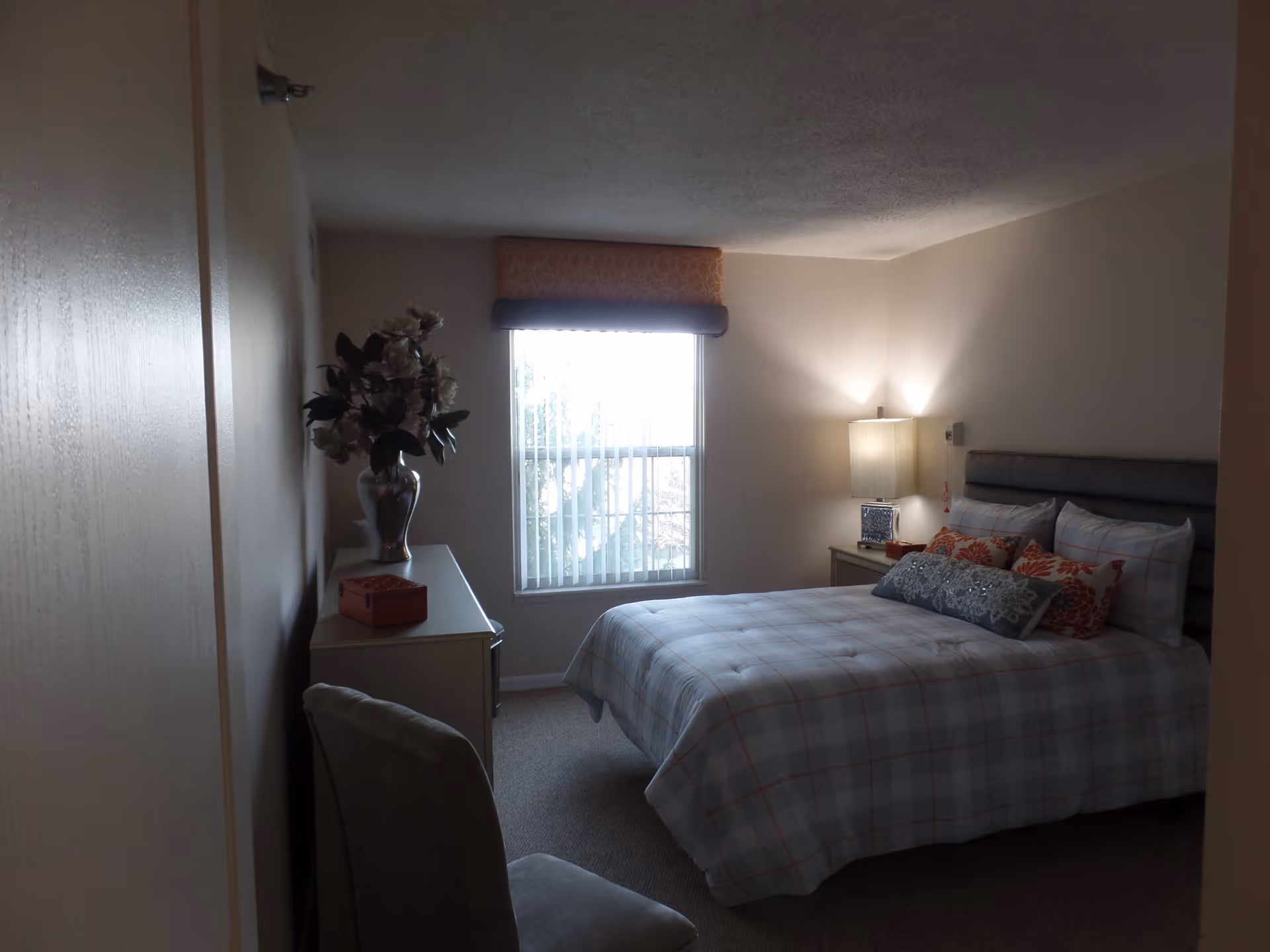 A bedroom with a bed covered in a plaid comforter and multiple pillows, a nightstand with a lamp, a window with vertical blinds, a dresser with a vase of flowers and a small box, and a chair in the foreground.