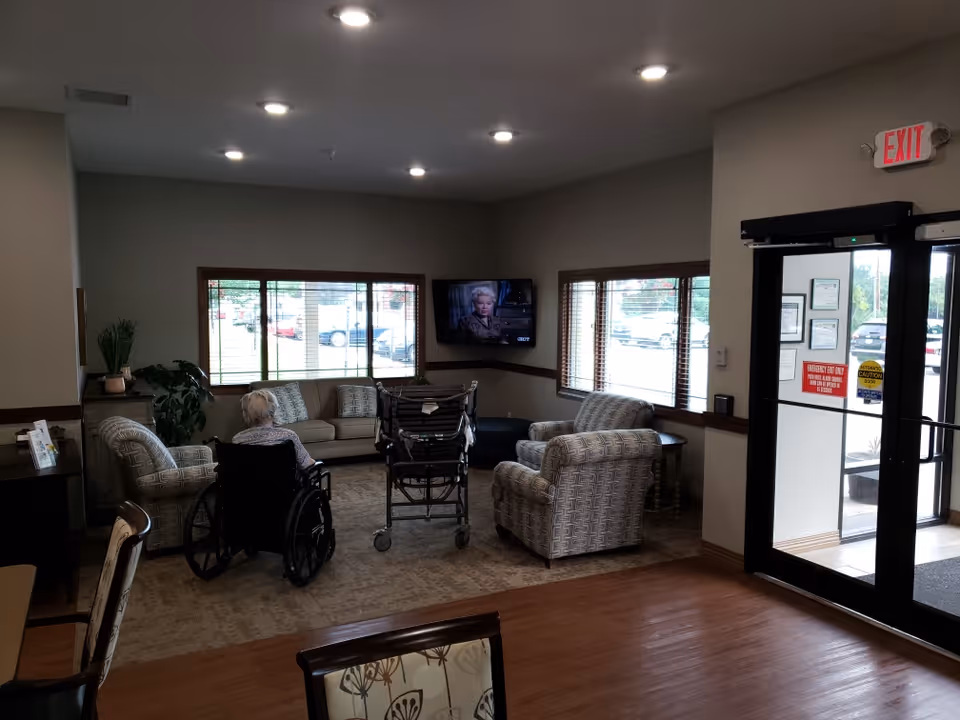 A common area in a senior living facility with a woman in a wheelchair watching a wall-mounted TV. The room has several cushioned chairs and a sofa, large windows with blinds, a wooden floor, and an exit door with signs.
