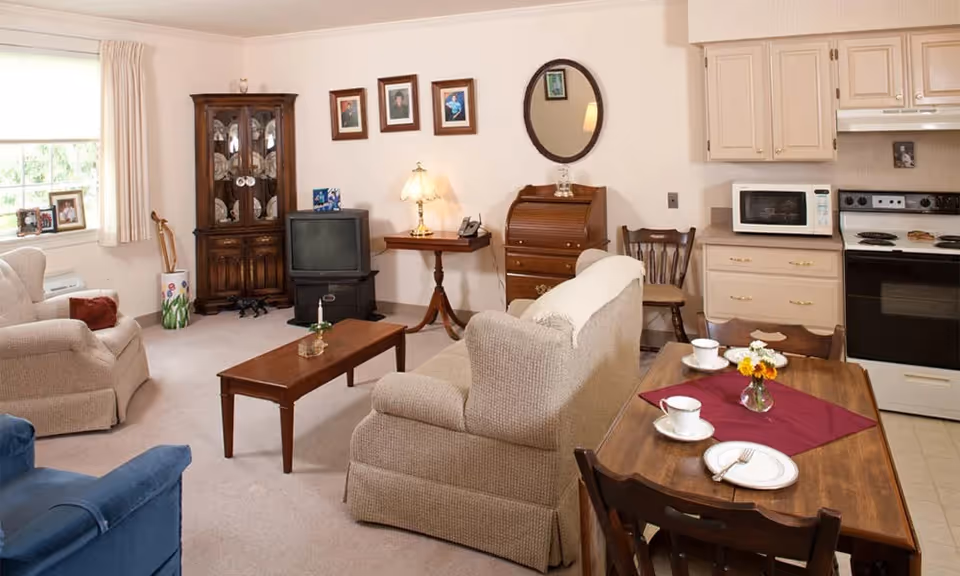 A cozy living area in a senior living facility featuring beige upholstered armchairs and sofa, a wooden coffee table, a small dining table set with cups and plates, a vintage TV on a stand, a wooden cabinet with glass doors, and a compact kitchen area with a stove, microwave, and cabinets. The walls are decorated with framed photos and a round mirror.