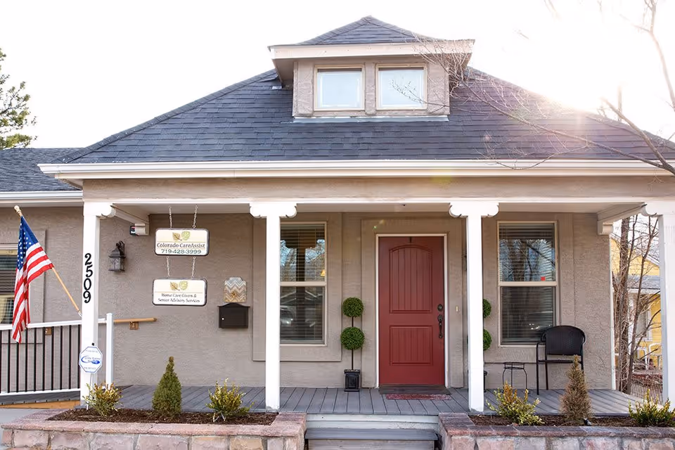 Front exterior view of a single-story building with a gray roof and beige walls. The entrance features a red door centered between two windows with white trim. There is a small porch with white columns, two topiary plants on either side of the door, a black chair and small table on the right side, and an American flag mounted on the left side. Two signs hang near the door, one reading 'Colorado CareAssist 719-428-3999' and the other 'Home Care Givers & Senior Advisory Services'. The building number 2509 is displayed vertically on the left porch column.