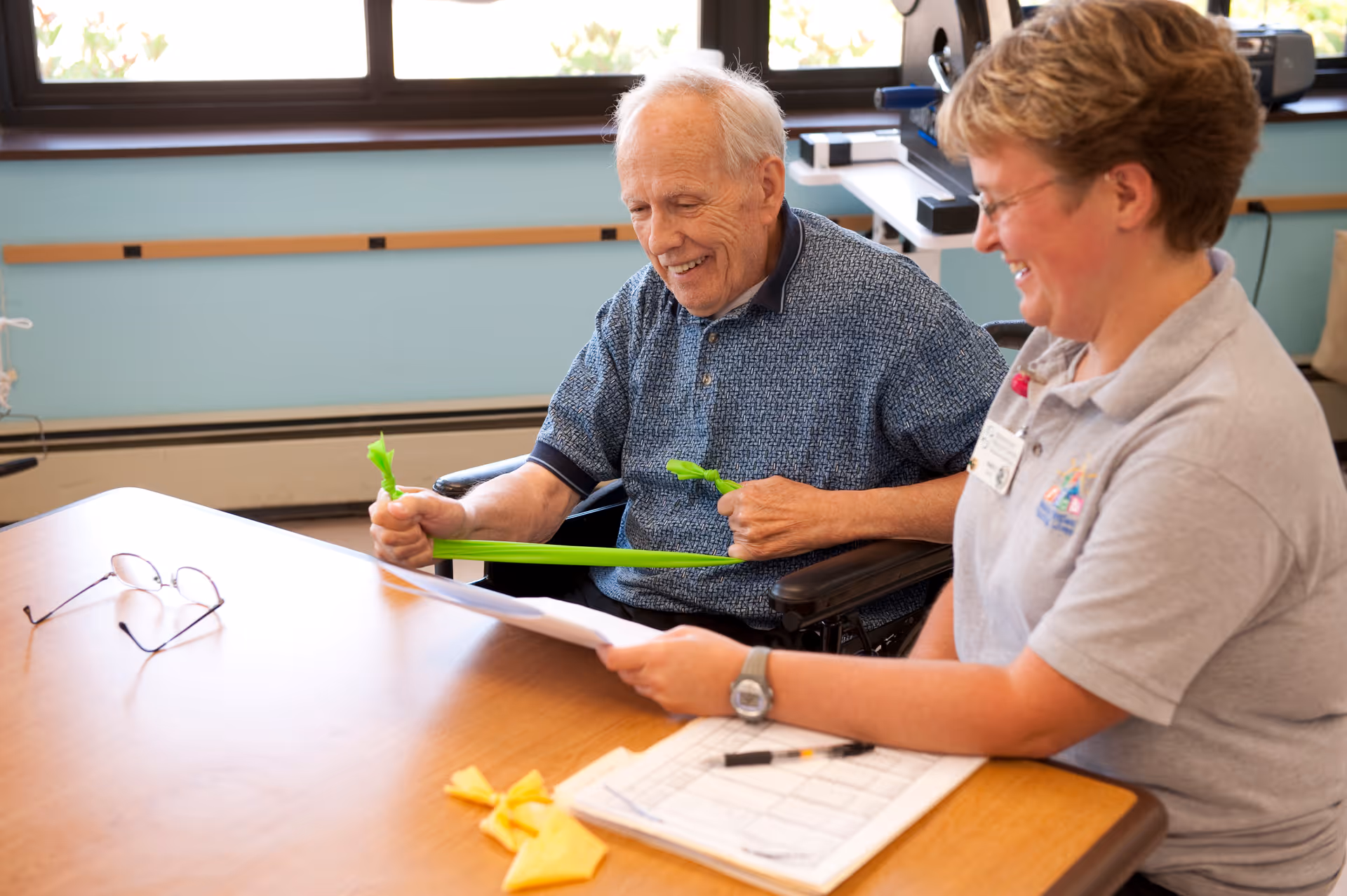 An elderly man in a wheelchair is exercising with a green resistance band while a smiling caregiver or therapist sits beside him holding a paper and a clipboard on a wooden table in a bright room with large windows.
