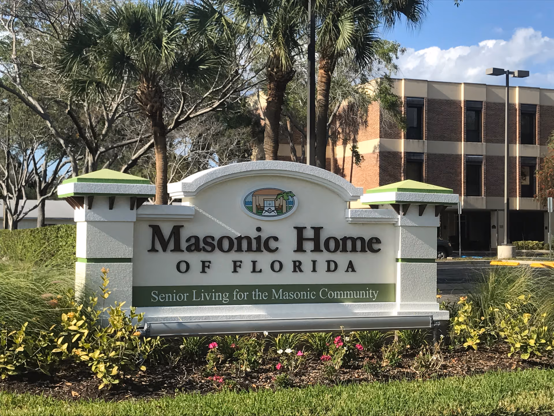 Outdoor view of a sign for Masonic Home of Florida, a senior living community. The sign is white with green accents and surrounded by landscaping with flowers and bushes. Behind the sign, there are palm trees and a multi-story brick building under a partly cloudy sky.
