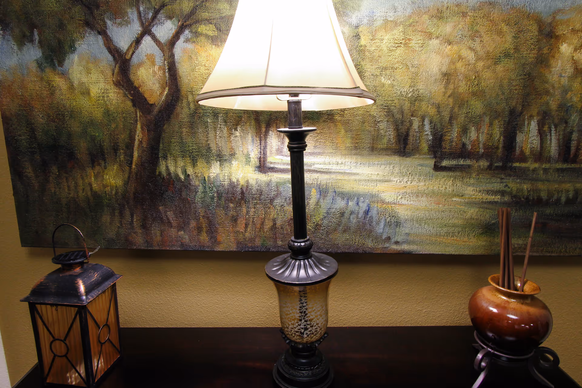 A decorative table setup featuring a lit table lamp with a beige shade, a rustic lantern on the left, and a brown ceramic vase with reed sticks on the right. Behind the table is a large painting depicting a forest scene with trees and greenery.