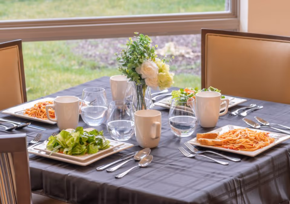 A dining table set for four with plates of salad and spaghetti with bread, white mugs, water glasses, silverware, and a small vase with white and green flowers. The table is covered with a dark tablecloth and is positioned near a window showing a grassy outdoor area.