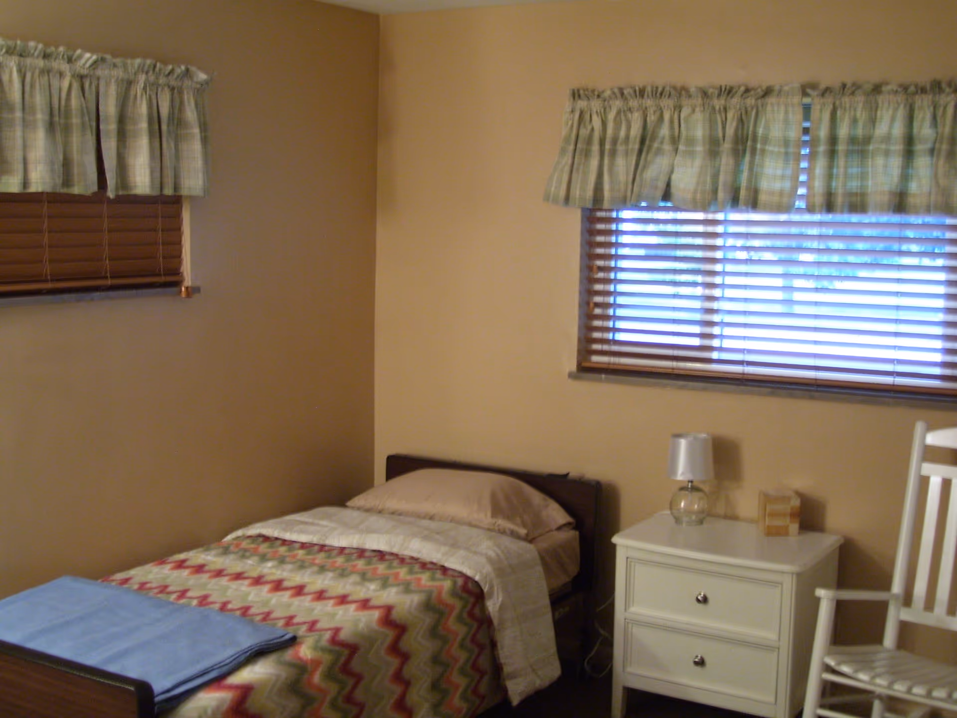 A small bedroom with a single bed covered with a colorful zigzag patterned blanket and a blue folded blanket at the foot. There are two windows with wooden blinds and plaid valances. Next to the bed is a white nightstand with a small lamp and a box of tissues. A white wooden rocking chair is partially visible on the right side.