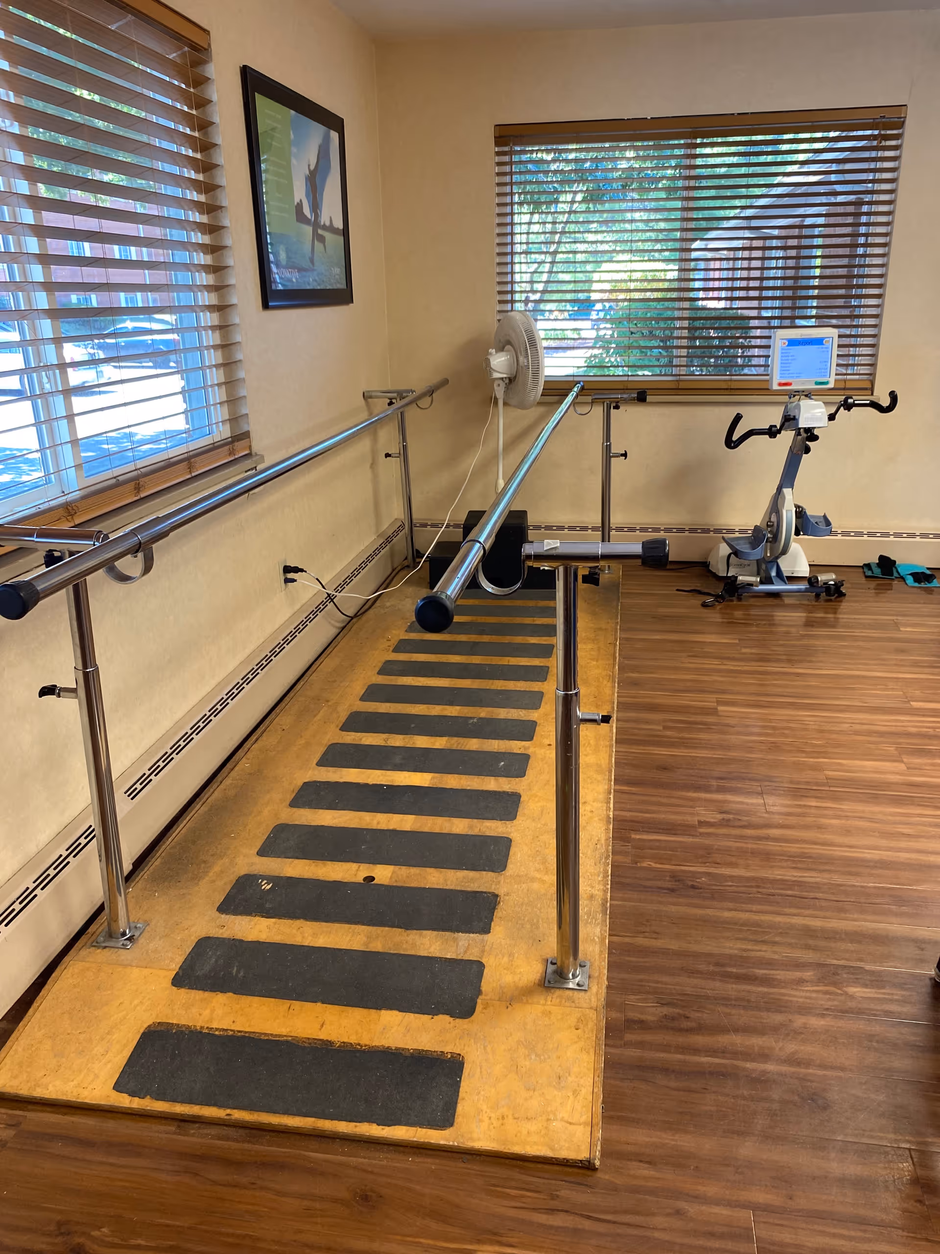 A physical therapy room with parallel bars for walking exercises on a wooden platform with black grip strips. There is a stationary exercise bike near a window with blinds, and a fan mounted on the wall. The room has wood flooring and beige walls with a framed picture hanging.