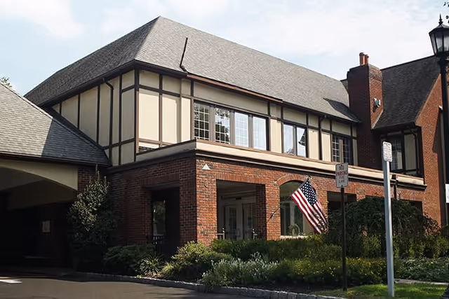 Exterior view of a two-story brick and beige building with a pitched roof, featuring multiple windows and an American flag near the entrance, surrounded by greenery and a street lamp.
