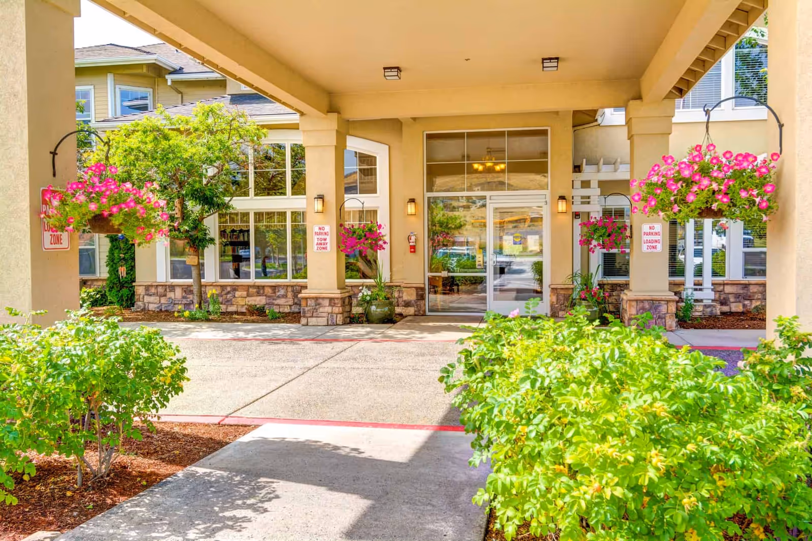Covered entrance of a residential facility with a porte-cochere, hanging flower baskets, glass double doors, and landscaped shrubs.