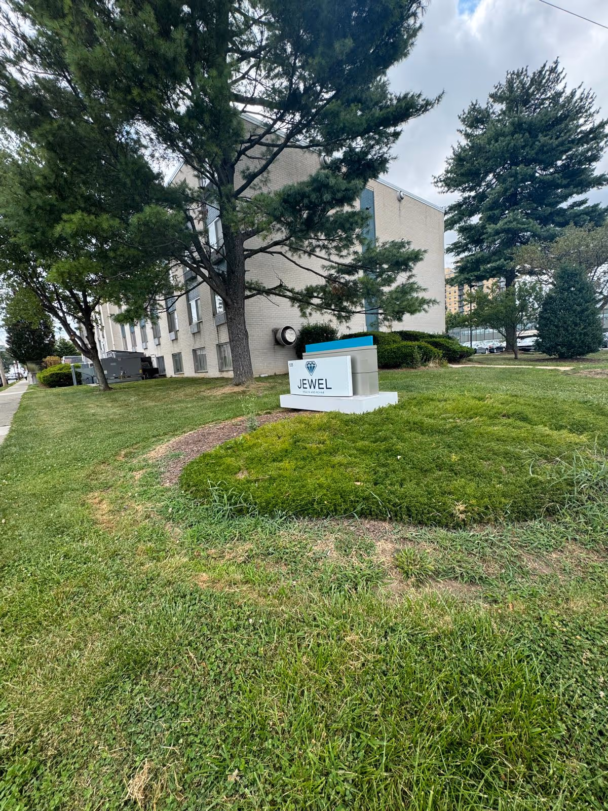 A low-rise brick building set behind trees and a grassy lawn with a ground-level sign reading "JEWEL".