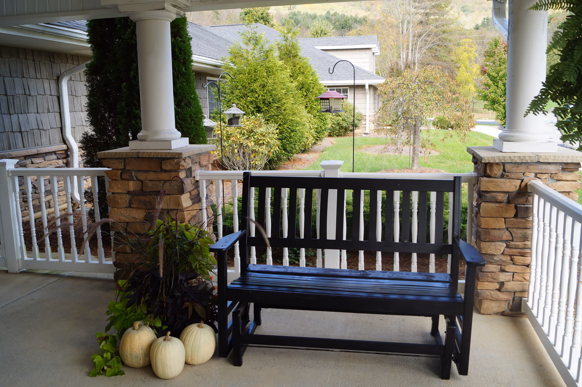A black wooden bench on a covered porch with stone pillars and white railings. Three white pumpkins and some green plants are placed on the ground next to the bench. In the background, there are green bushes, a tree, and parts of a building with a gray roof.