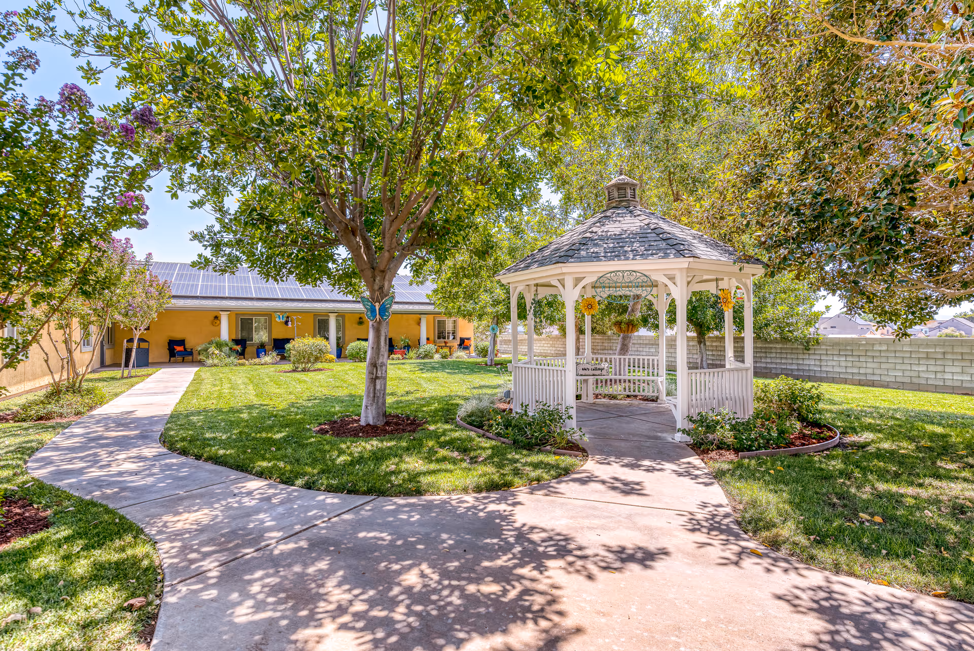 Sunny courtyard with a white gazebo, paved walkways, trees, and a single-story building in the background.