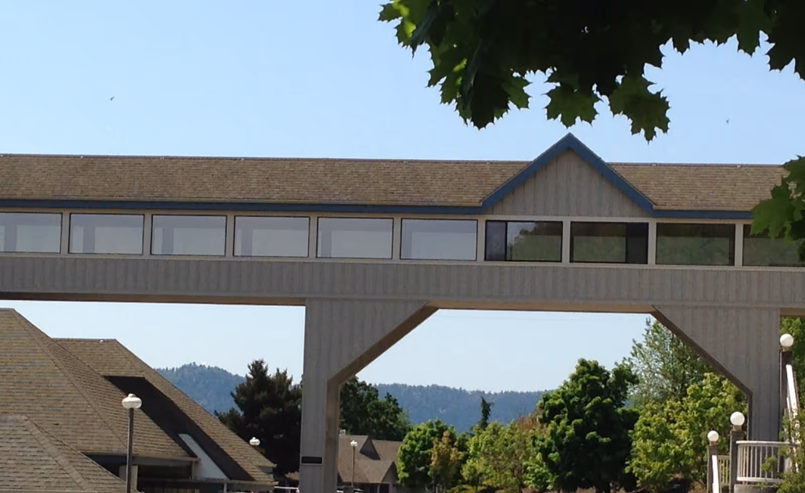 Exterior view of a building at Linus Oakes Village featuring a covered elevated walkway with multiple windows, surrounded by trees and other buildings under a clear blue sky.