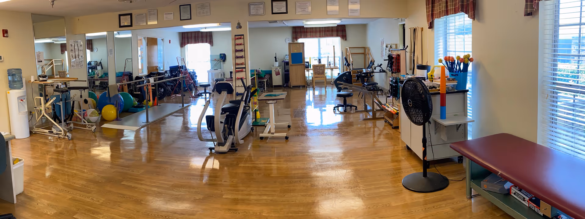 A spacious physical therapy or rehabilitation room with wooden floors, exercise equipment including stationary bikes, therapy balls, parallel bars, and various rehabilitation tools. The room has large windows with blinds and curtains, mirrors on one wall, and certificates or awards displayed above the mirrors.