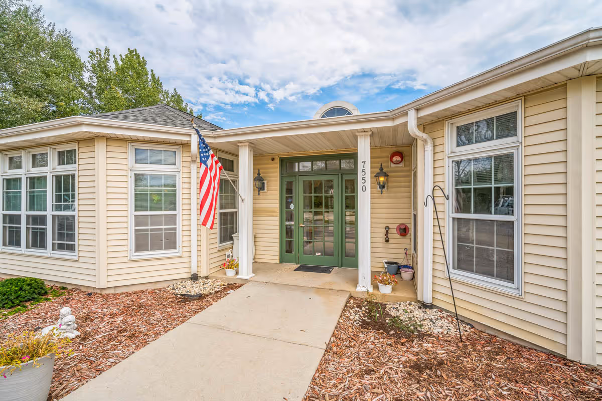 Front entrance of a beige single-story building with green double doors, an American flag mounted on a pole, two white columns, and several windows. The building number 7550 is displayed on one of the columns. There are small flower pots and landscaping with mulch and rocks around the entrance.
