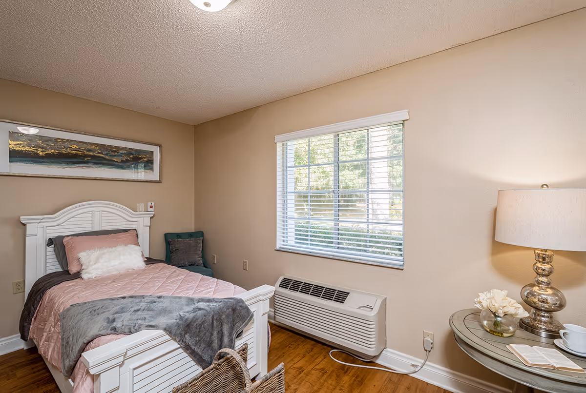 A cozy bedroom with a single white wooden bed featuring pink and gray bedding. There is a small green chair in the corner, a window with white blinds letting in natural light, and a round side table with a lamp, a small flower arrangement, and an open book. The walls are beige and the floor is wooden.