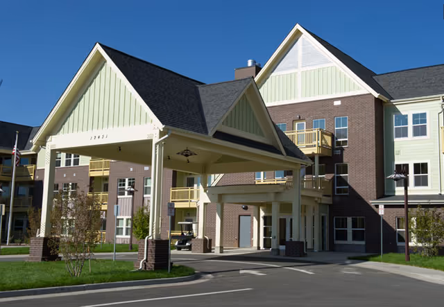 Exterior view of The Avenues at Crofton Park senior living facility showing a covered entrance with a peaked roof supported by columns, a driveway, and a multi-story building with balconies and windows in the background.