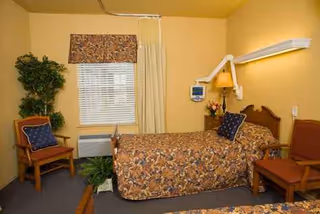 Bedroom in a senior facility with floral quilt, window, plants, chairs, and bedside lamp.