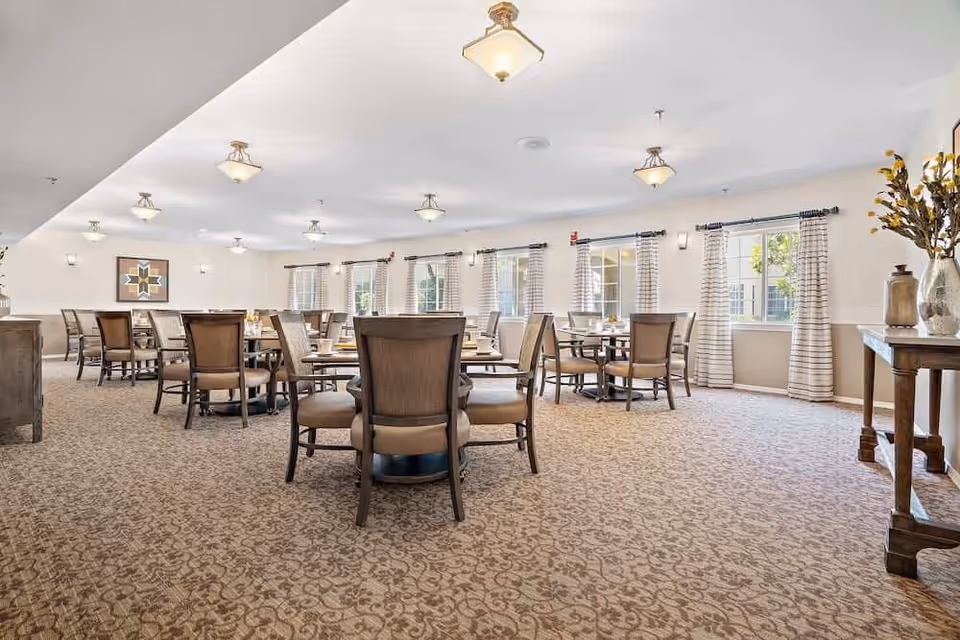 A spacious dining room with multiple round tables and cushioned chairs arranged neatly. The room has large windows with striped curtains allowing natural light to fill the space. The carpet has a patterned design, and the walls are decorated with framed artwork and a console table with vases and decorative items. Ceiling lights provide additional illumination.