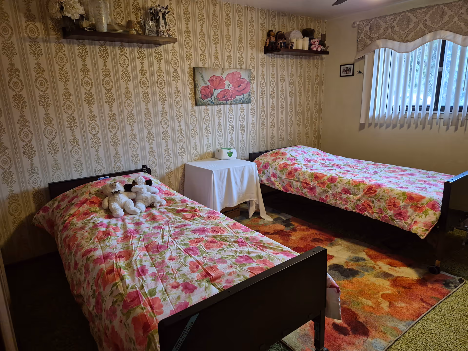 A cozy bedroom with two single beds covered in floral bedspreads featuring pink and red flowers. Each bed has a stuffed animal placed on top. The room has patterned wallpaper with a vintage design, two wooden shelves holding decorative items, a small table covered with a white cloth between the beds, and a window with vertical blinds and a valance. A colorful rug is on the floor beneath the beds.