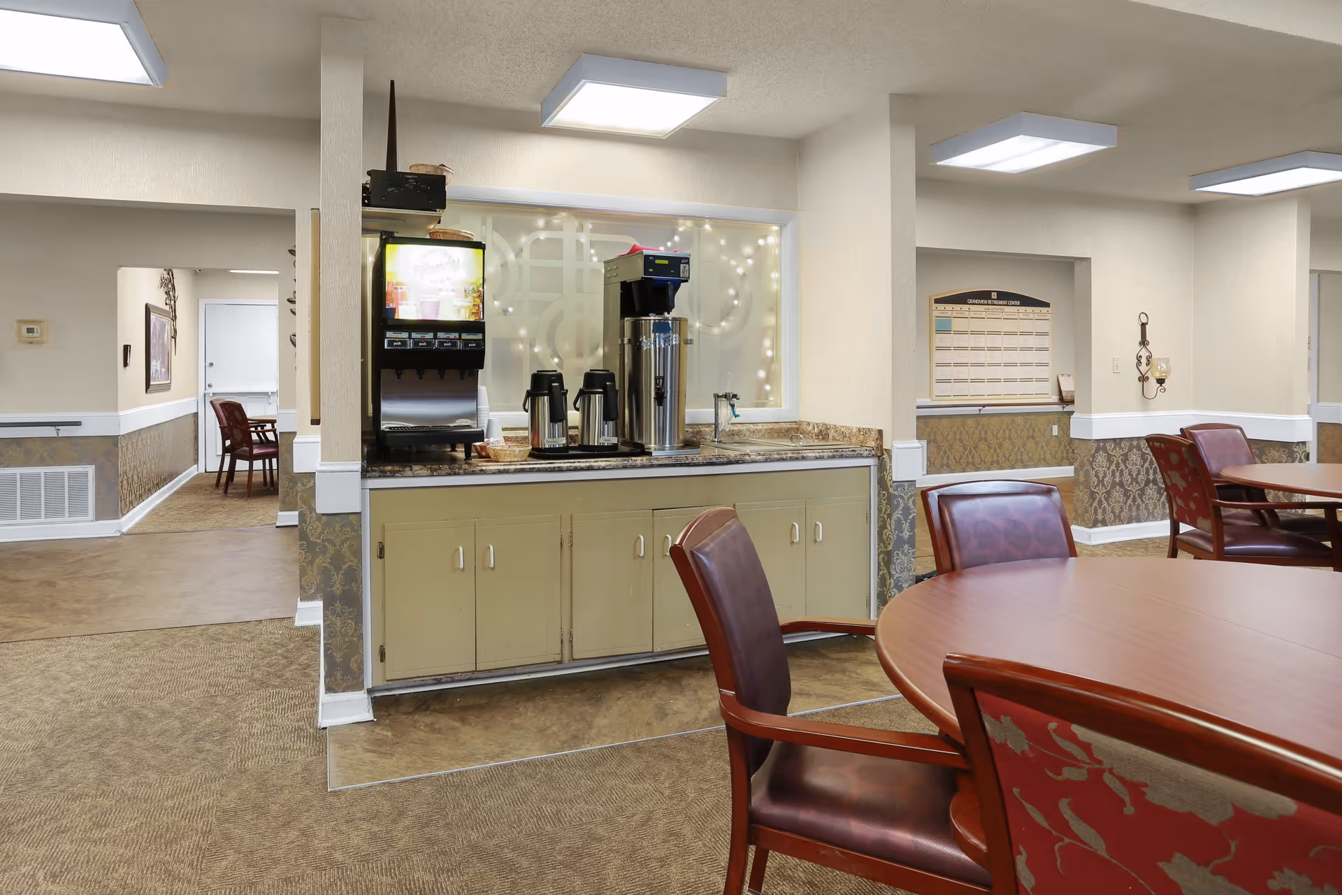 Interior view of a common area in a senior living facility with a beverage station featuring a coffee machine, dispensers, and coffee pots on a countertop. There are round wooden tables with red cushioned chairs around them. The walls have a beige and patterned wallpaper with white trim, and the floor is carpeted. A hallway with additional seating is visible in the background.
