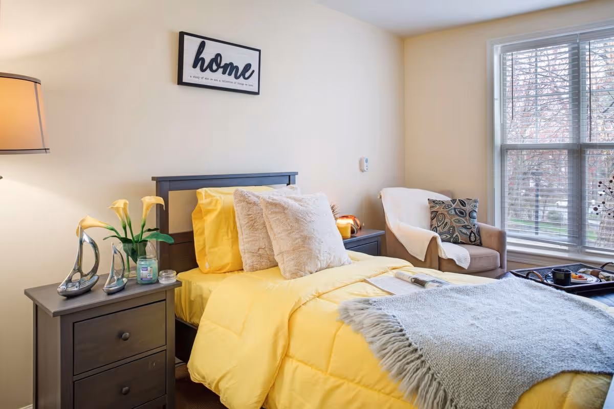 Well-lit bedroom with a single bed in yellow bedding, bedside tables, a cozy armchair by a large window, and a framed 'home' sign on the wall.