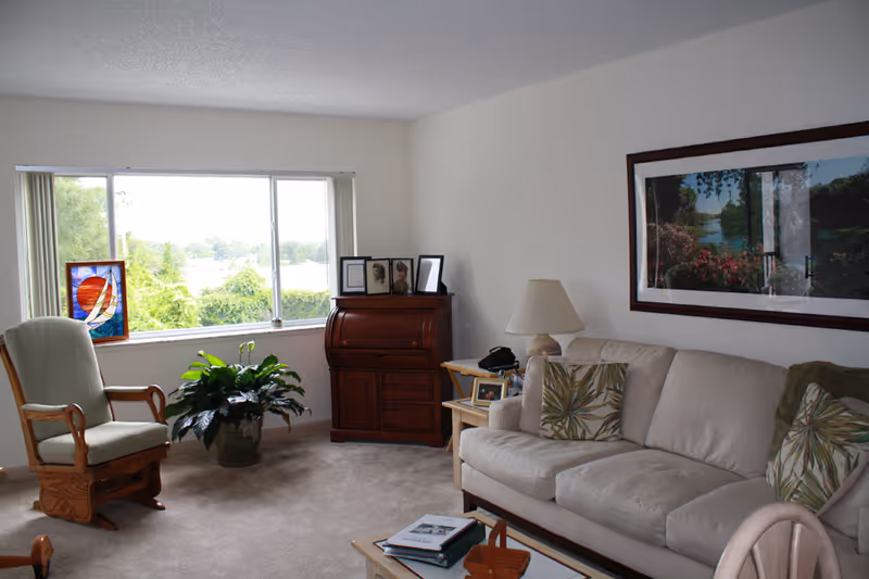 A cozy living room with a beige couch adorned with green and beige patterned pillows, a wooden rocking chair with light green cushions, a wooden roll-top desk with framed photos on top, a side table with a lamp and a telephone, a large window showing greenery outside, a potted plant near the window, and a framed landscape painting on the wall.
