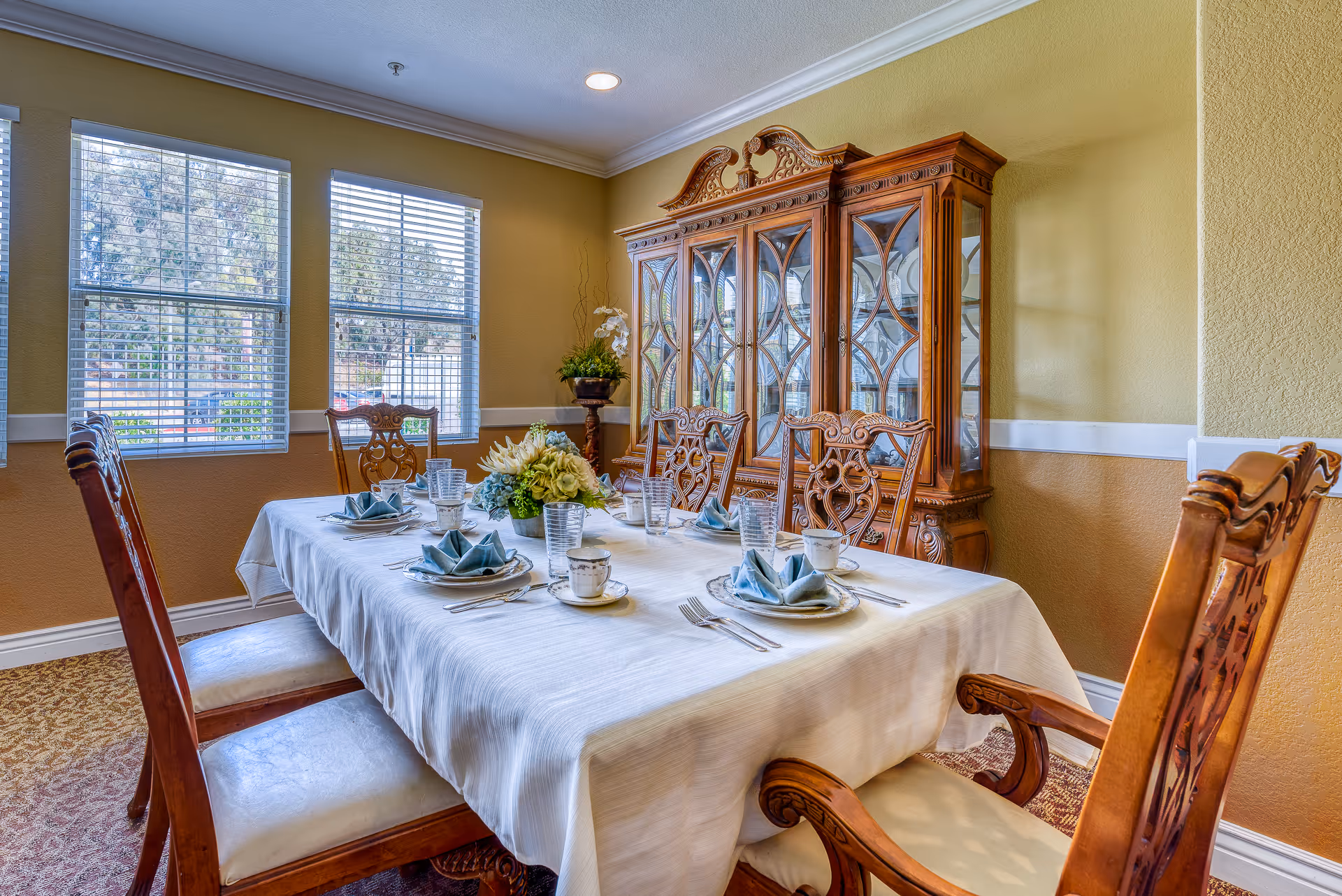 A formal dining room with a long table covered with a white tablecloth, set with plates, cups, glasses, and folded blue napkins. The room has wooden chairs with intricate designs, a large wooden china cabinet, and three windows with blinds letting in natural light.