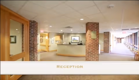 Reception area inside Tudor Oaks Senior Living Community featuring a curved reception desk, brick pillars, beige walls, and a hallway leading to other rooms.