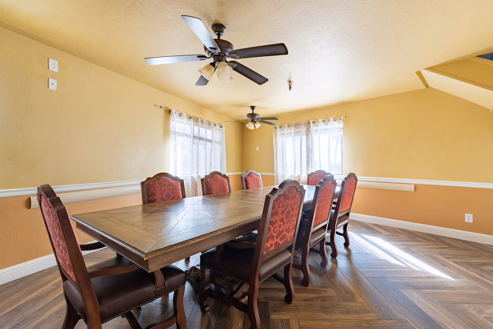 Bright dining room with a long wooden table surrounded by red-upholstered chairs, ceiling fans, and windows with sheer curtains.