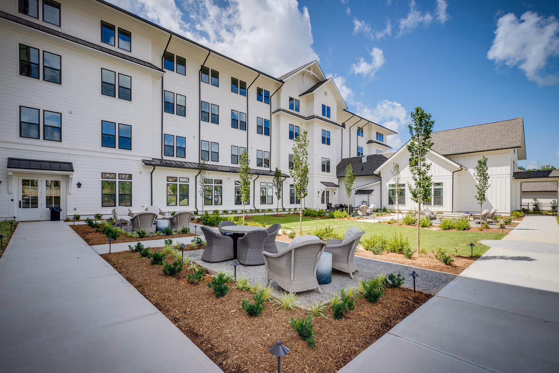 Outdoor courtyard area of a senior living facility with white multi-story building in the background, featuring several seating areas with wicker chairs and tables surrounded by landscaped garden beds and young trees under a partly cloudy blue sky.