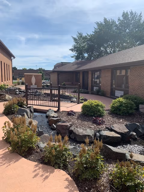 Outdoor courtyard with a small bridge over a rock-lined water feature, landscaped beds, and surrounding brick buildings.