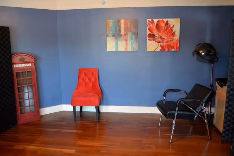 Empty interior room with a red upholstered chair, a black salon chair under a hair dryer, colorful wall art on a blue wall, and a red phone booth prop.