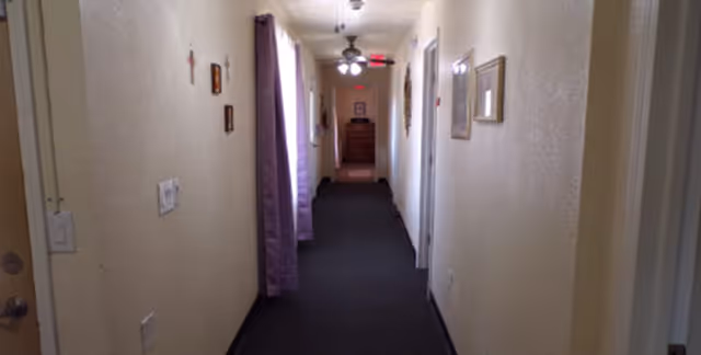 Carpeted interior hallway in a senior living facility with doors on both sides, wall art, purple curtains and a dresser at the far end.