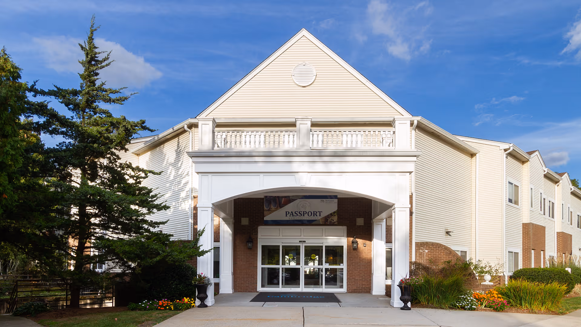 Front exterior view of a two-story building with beige siding and brick accents, featuring a covered entrance with white columns and a sign above the door that reads 'PASSPORT'. There are trees and landscaped bushes with flowers on either side of the entrance under a blue sky with some clouds.