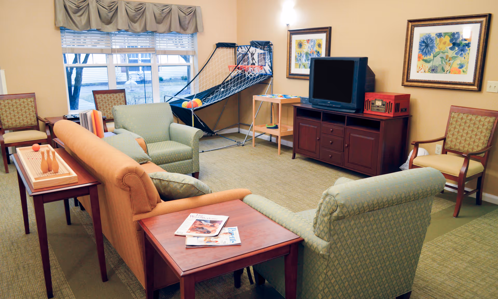 A cozy common room with green and orange upholstered chairs and a sofa arranged around a wooden TV stand holding an old-style television and a red radio. The room features a basketball arcade game, a small wooden table with a ring toss game, framed floral artwork on the walls, and large windows with beige curtains letting in natural light.