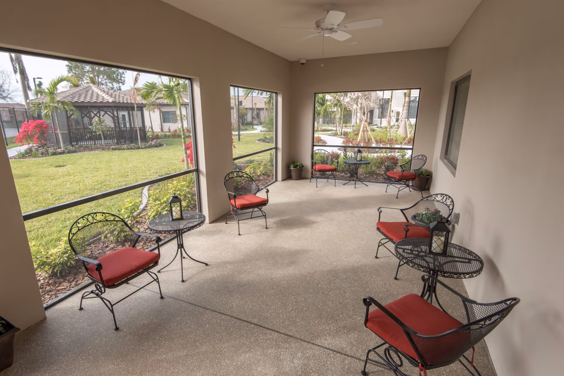 A screened-in patio area with six black metal chairs featuring red cushions and three matching round tables. The patio overlooks a landscaped garden with green grass, palm trees, flowering plants, and a gazebo in the background.
