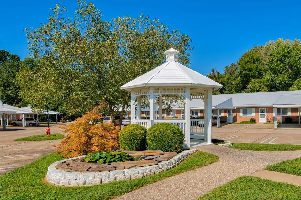 White gazebo in a landscaped courtyard with trees, shrubs, and a parking lot nearby.