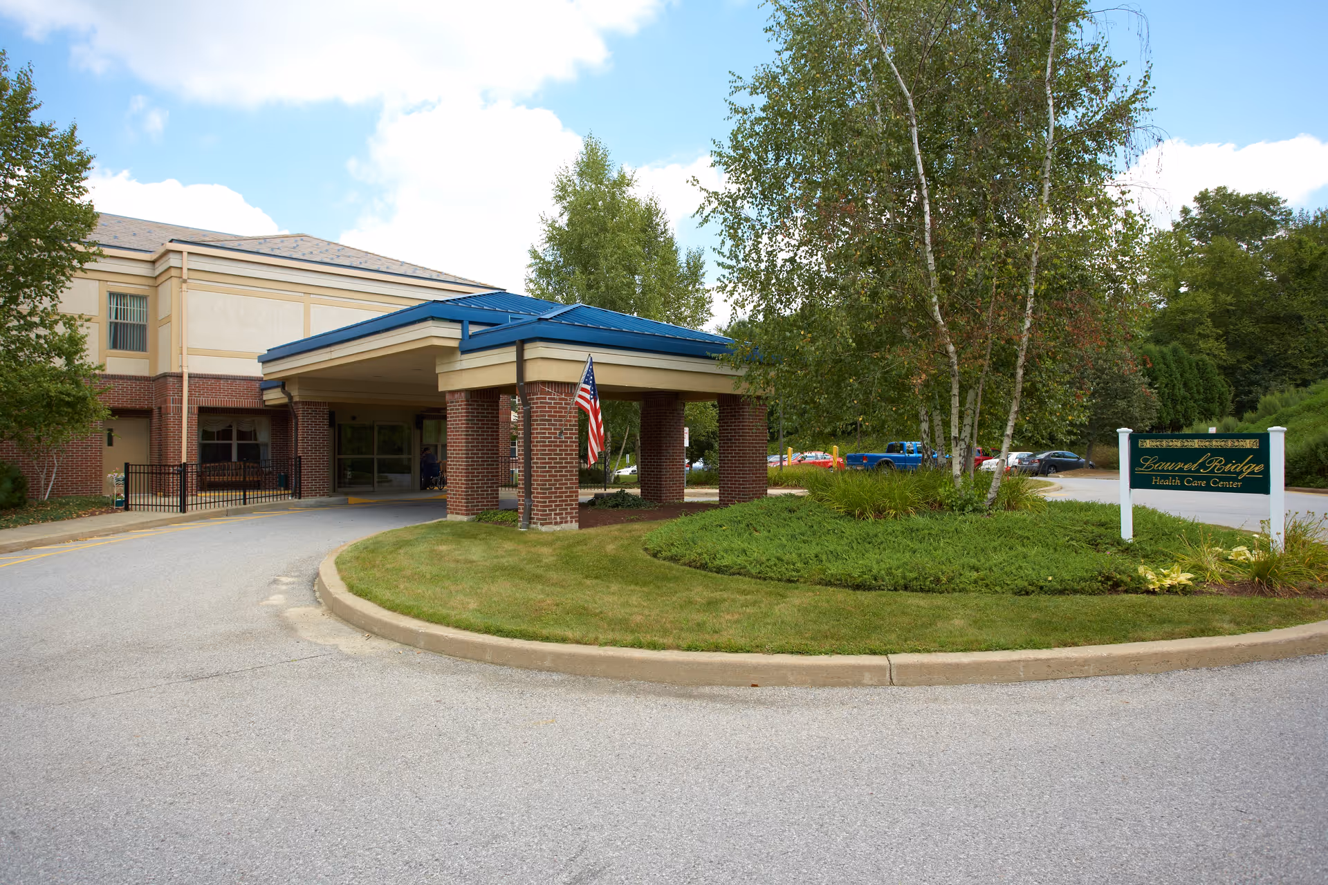 Exterior view of Laurel Ridge Health Care Center showing the main entrance with a covered drop-off area supported by brick columns, an American flag hanging from one column, a landscaped roundabout with green grass and trees, and a sign displaying the facility's name.