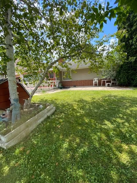 A sunny outdoor garden area with green grass, a tree with leafy branches, a small raised garden bed with a statue, and a building with a brick and siding exterior. There are two white chairs and a small table on a concrete patio near the building.