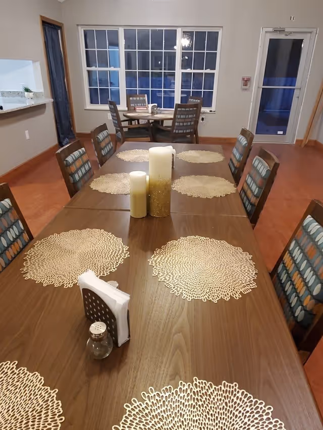 Interior view of a dining room with a long wooden table set with decorative placemats, three pillar candles in the center, a napkin holder, and a salt shaker. There are cushioned chairs around the table and a smaller round table with chairs near large windows in the background. A door with a glass panel is visible on the right side.