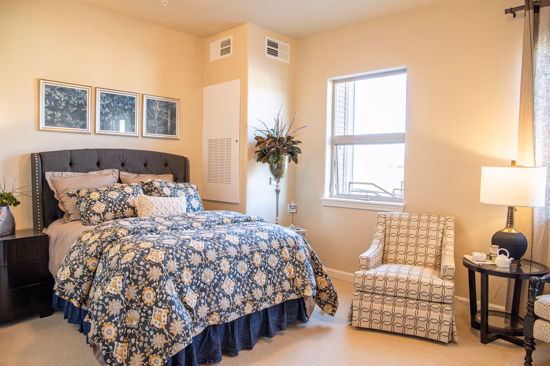 A cozy bedroom with a bed featuring a dark upholstered headboard and floral patterned bedding in shades of blue, beige, and white. There are three framed botanical prints on the wall above the bed. To the right of the bed is a window letting in natural light, a patterned armchair, and a round side table with a lamp and tea set. A tall vase with decorative plants is placed in the corner near the window.