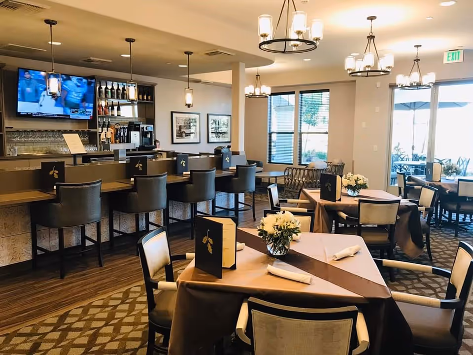 Well-lit dining room with tables set with menus and floral centerpieces, a bar with high stools and a wall-mounted TV in the background.