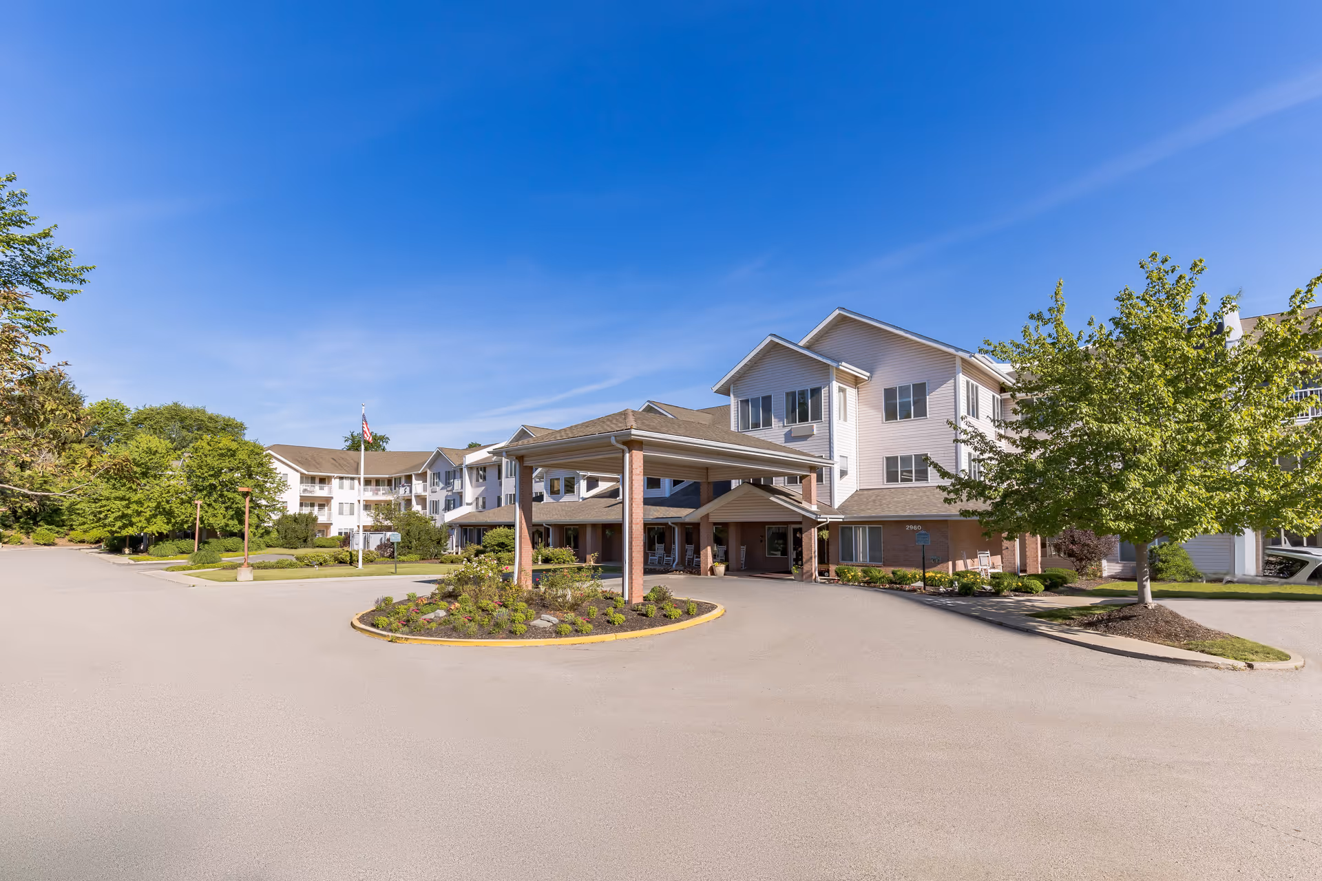 Exterior view of a multi-story senior living facility building with a covered entrance, surrounded by trees and landscaping under a clear blue sky.
