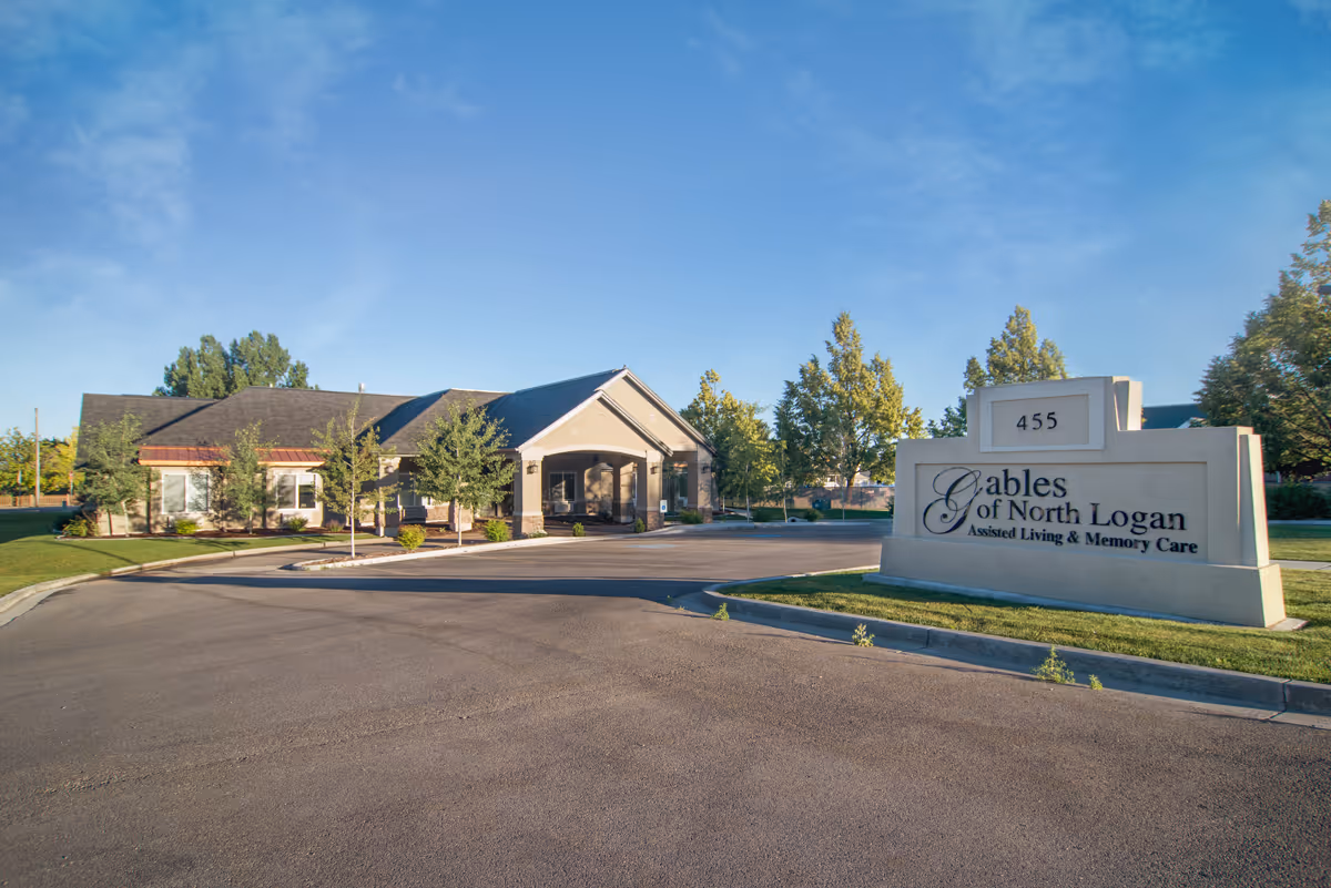 Exterior view of The Gables Assisted Living of North Logan building with a large sign displaying the facility name and address number 455, surrounded by trees and a clear blue sky.