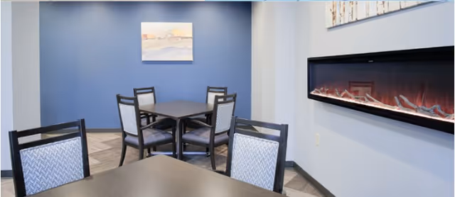 A cozy indoor seating area with dark wooden tables and chairs featuring patterned cushions. The room has a blue accent wall with a framed landscape painting and a modern electric fireplace mounted on a light gray wall.